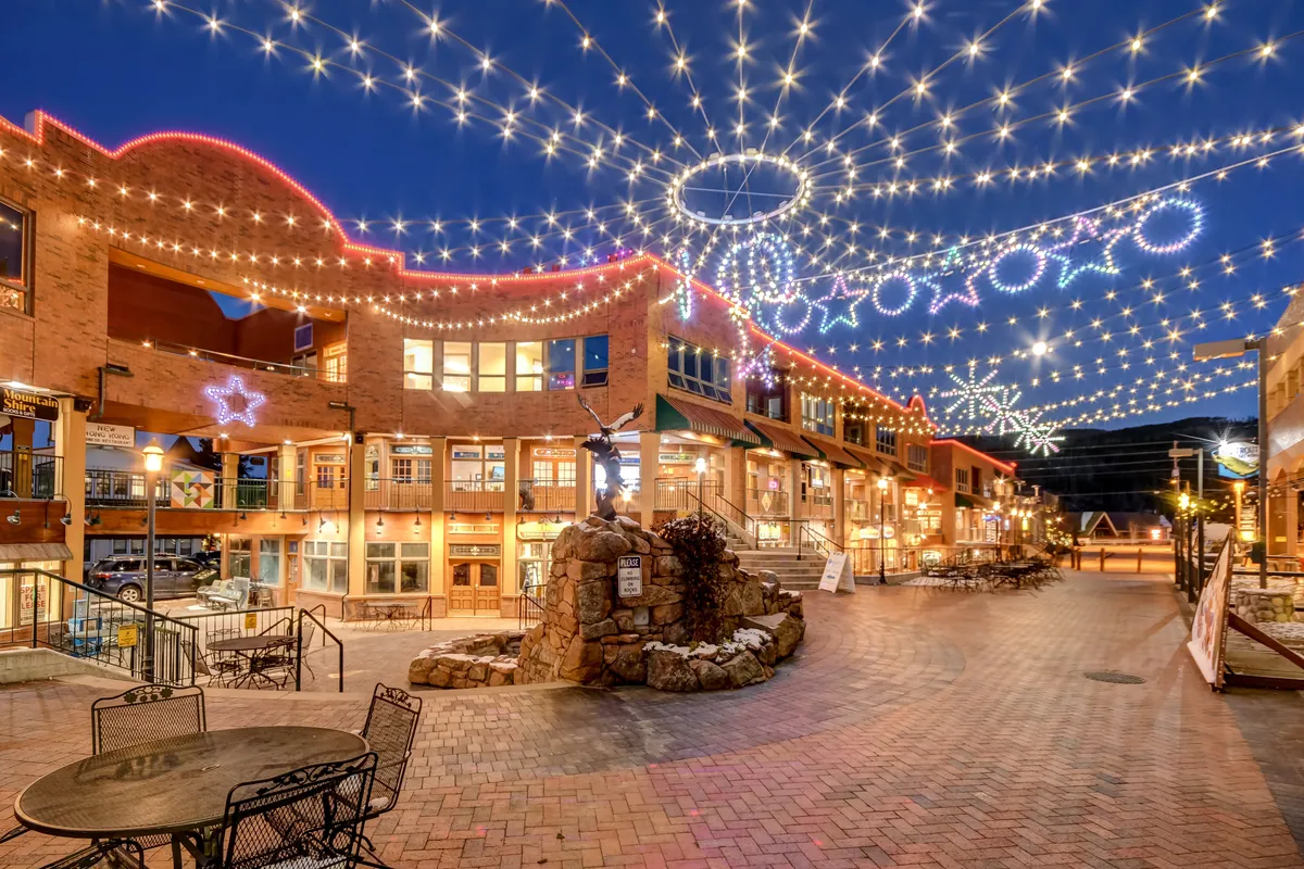An inviting outdoor plaza scene, with decorative string lights overhead, a stone fountain, comfortable seating, and buildings decorated for the season. People are seen dining, shopping, and socializing, capturing the lively energy of Winter Park’s downtown nightlife and après-ski scene.