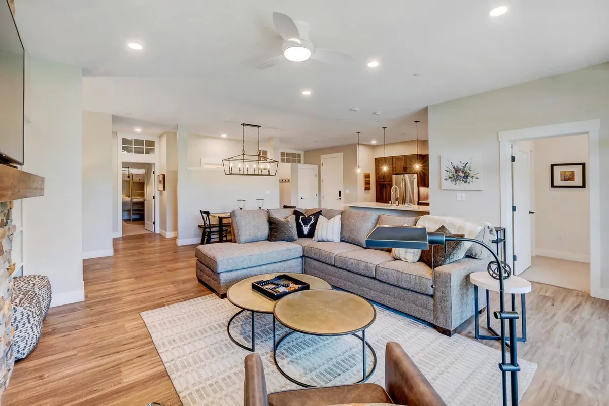 The image presents a wide-angle view of an open-plan living area that seamlessly connects a large sectional sofa, coffee table, dining area, and adjacent kitchen. Natural light streams through generous windows, and the overall design showcases contemporary mountain decor, creating a warm and welcoming atmosphere ideal for family relaxation and entertaining.