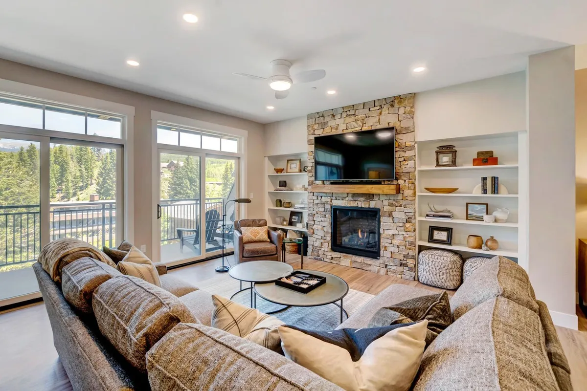 A spacious, light-filled living room featuring a plush sectional sofa centered on a modern fireplace and large TV, flanked by stylish bookshelves. Generous windows frame sweeping mountain views, inviting natural light and offering a cozy yet elegant space for relaxation, movie nights, or gathering with friends and family.