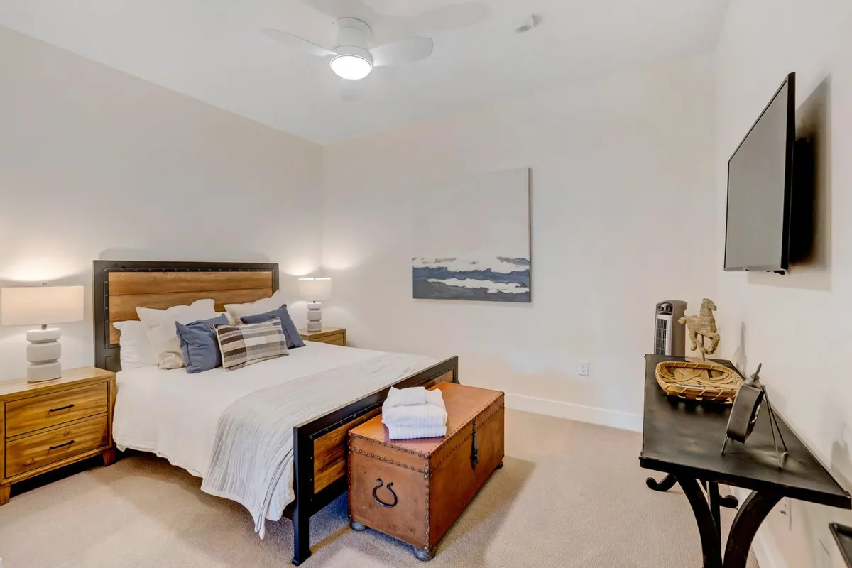A soothing queen bedroom with a comfortable bed adorned with decorative pillows and premium linens. Nightstands with lamps flank the bed, artwork adds a personal touch, and a vintage trunk sits at the foot of the bed.