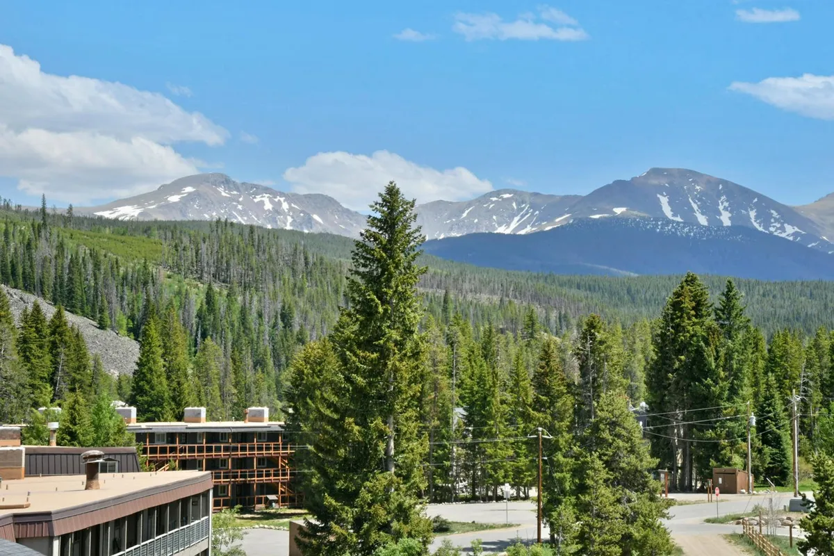 This exterior shot captures a modern building nestled against the backdrop of majestic mountains, surrounded by dense pine trees beneath a clear blue sky. The scene radiates tranquility and the promise of adventure, suggesting immediate access to nature and the outdoors.