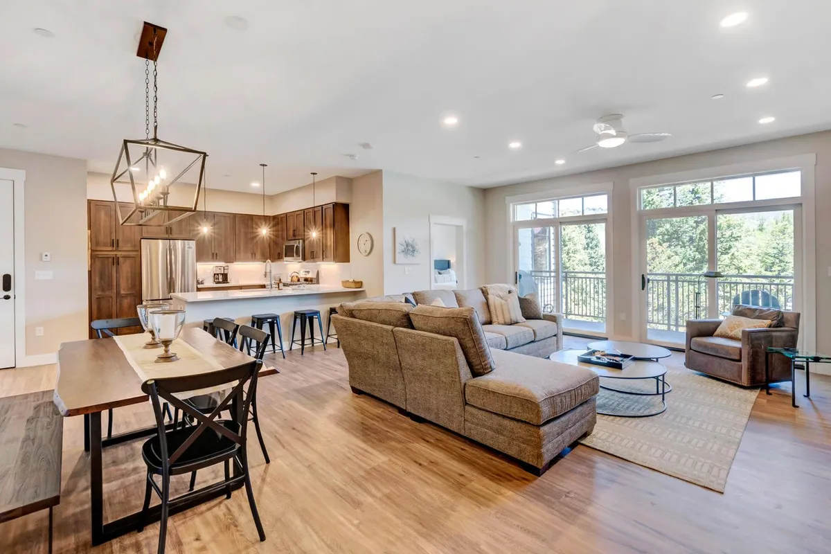 A cozy, open-plan living area featuring a plush sofa, a dining table set for casual meals, a sleek kitchen island, and large windows offering glimpses of the mountains outside. The space feels bright and welcoming, inviting guests to gather and unwind after a day of adventure.