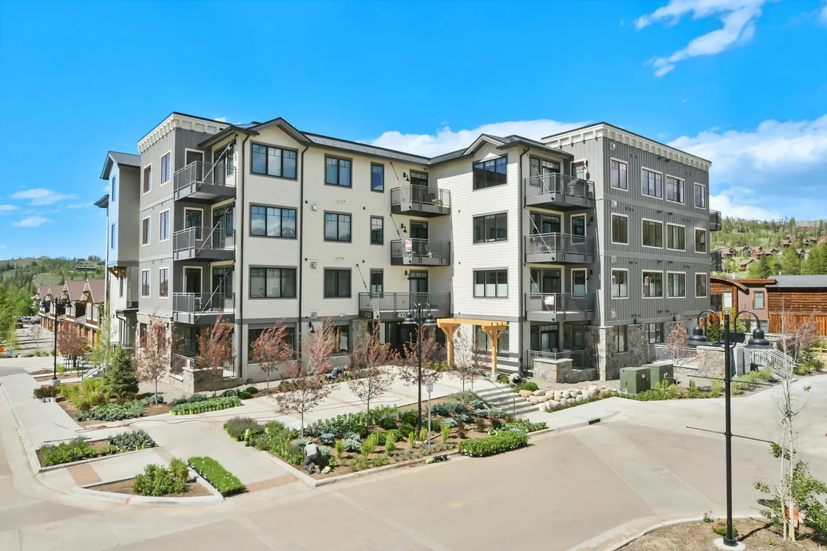 A welcoming four-story building with private balconies overlooks landscaped gardens and a meandering walkway, set beneath a vibrant sunset sky. Residents can be seen relaxing on balconies and strolling through the gardens, enjoying the communal and scenic atmosphere.