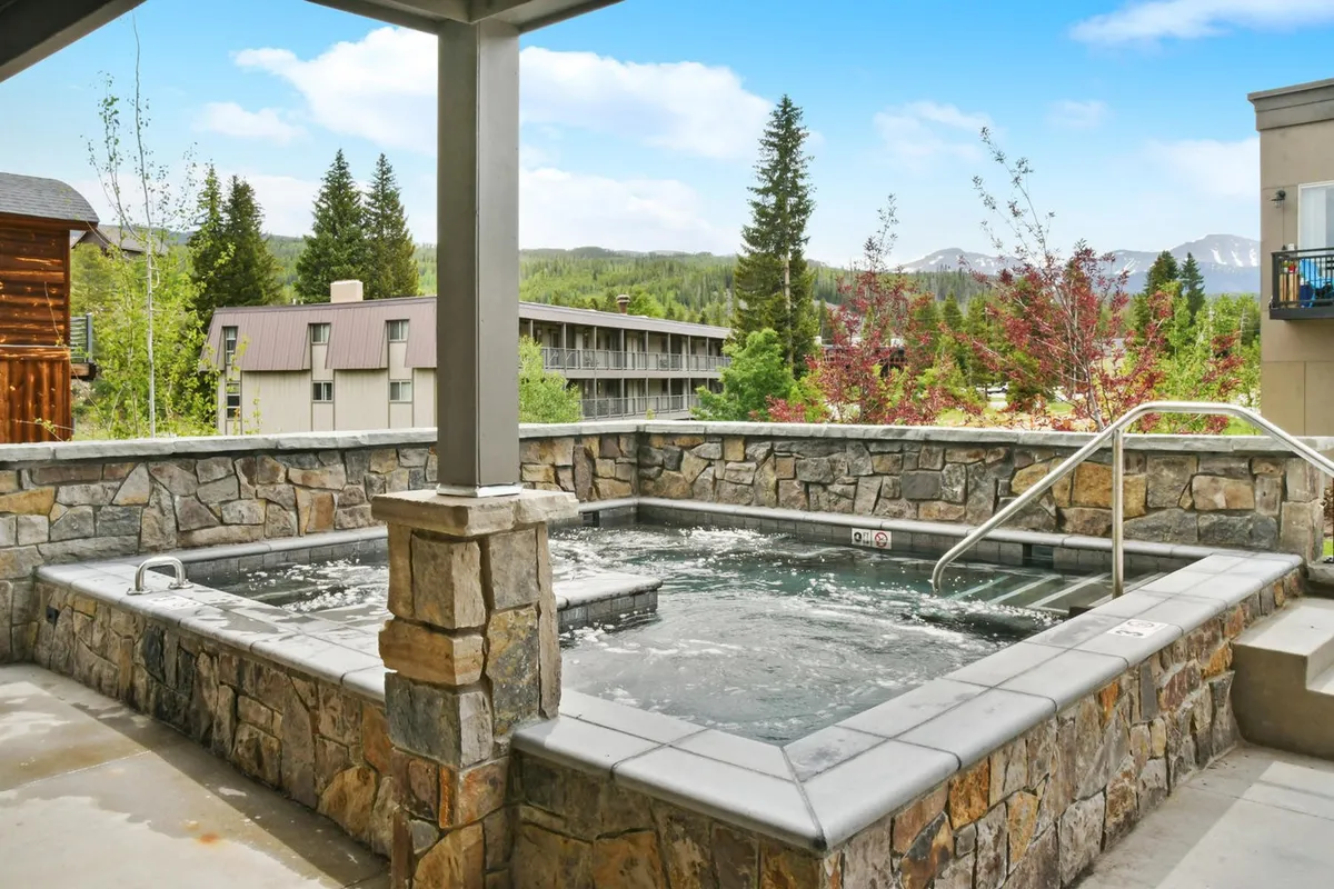 A large, stone-lined hot tub set against a backdrop of natural mountain landscape, with several people relaxing and socializing in the warm water as the sun sets. The setting conveys a serene, resort-like ambiance and offers starlit evenings and scenic mountain views.