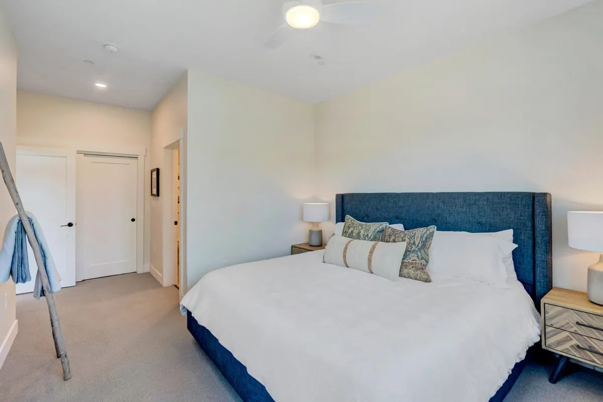 A serene bedroom with a large bed dressed in plush linens, matching nightstands, decorative pillows, and a ceiling fan above. The space is softly lit, creating a tranquil atmosphere suited for restful nights or relaxed mornings with coffee in bed.