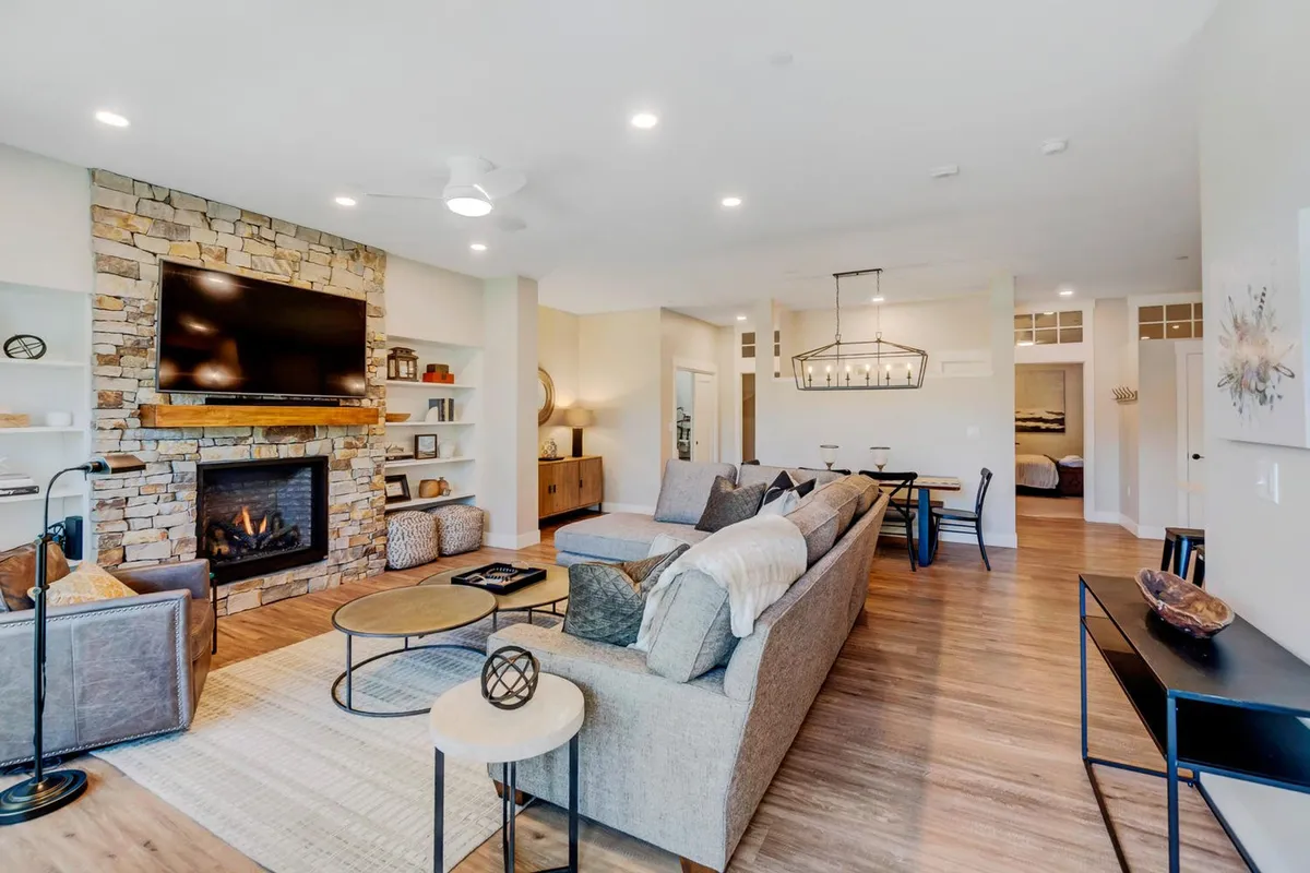 This image showcases a spacious, open-concept living room featuring a beautiful stone fireplace as its centerpiece. A large, comfortable sectional sofa and a coffee table create a perfect spot for family gatherings or board games. Floor-to-ceiling windows flood the space with natural light and offer breathtaking mountain views, making the room feel both luxurious and cozy.