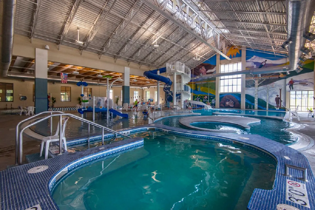An energetic indoor recreation scene featuring a lively swimming pool with water slides, children playing, and families relaxing poolside. The bright, playful atmosphere showcases family-friendly amenities available year-round at the nearby Grand Park Rec Center.