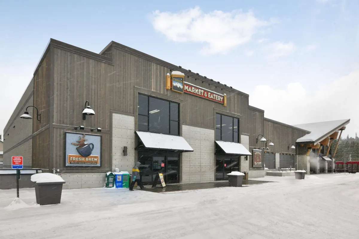 The image features the welcoming entrance to the Fireside Market, complete with a market sign, an inviting snow-covered exterior, and an outdoor seating area where people are seen shopping for groceries and enjoying hot beverages.