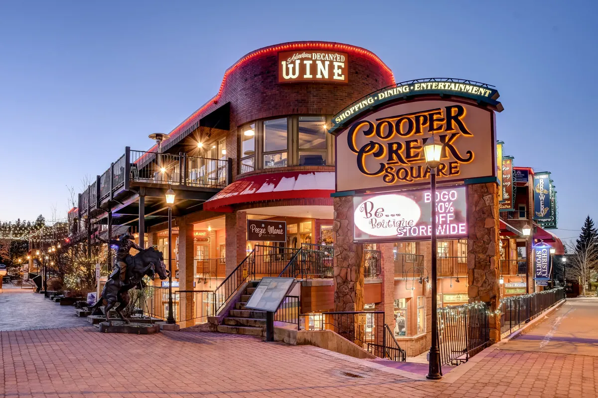 This exterior image captures the entrance to Cooper Creek Square, featuring a prominent sign, wine shop signage, a staircase, and a horse sculpture. The scene hints at a lively, festive environment perfect for family outings, shopping, and social gatherings in a walkable downtown area.